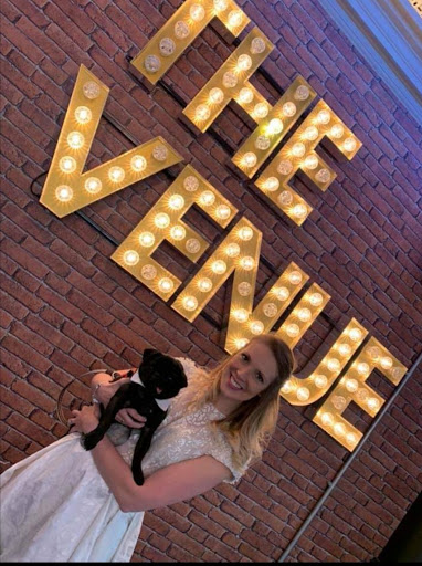 A female wedding chaperone holding a black pug stood in front of the words "The Venue" written in lightbulbs on a wall behind them