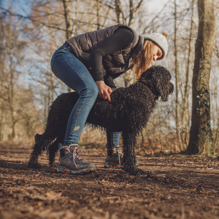 Muck Boots - A woman is crouched over a black dog. The woman is wearing a grey hat, a grey jacket, blue jeans and boots.