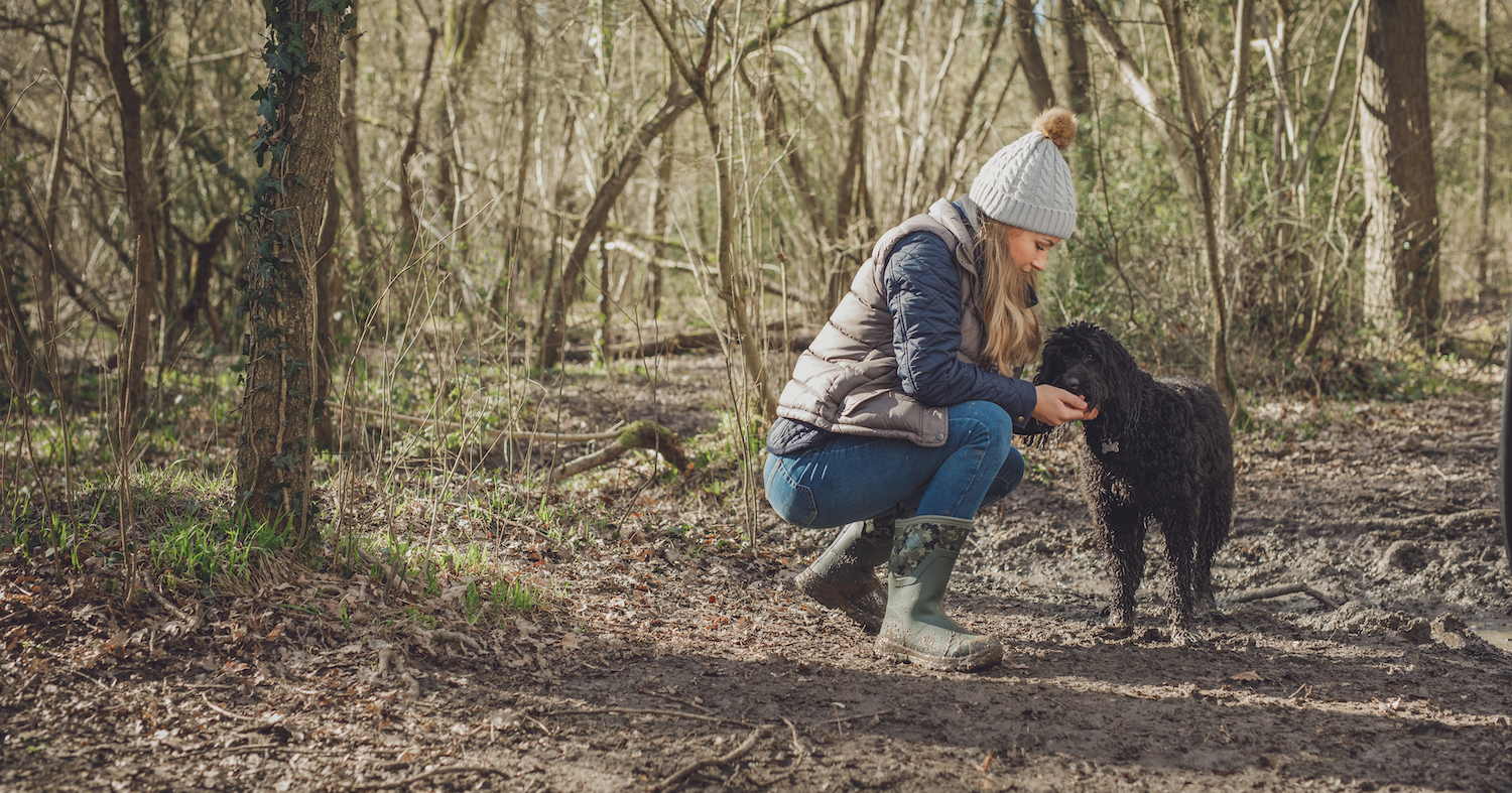 Muck Boots - A woman is crouched next to a black dog. The woman is wearing a grey hat, grey jacket, blue jeans and green boots.